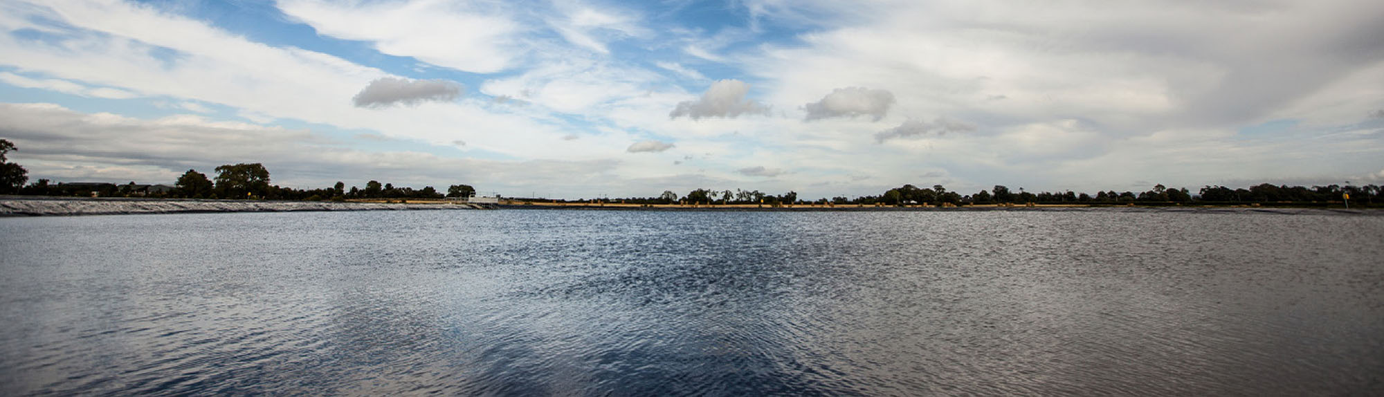 Wide panoramic view of a calm lake under a cloudy sky, with gentle ripples on the water, scattered trees along the distant shoreline, and patches of blue sky breaking through the clouds.