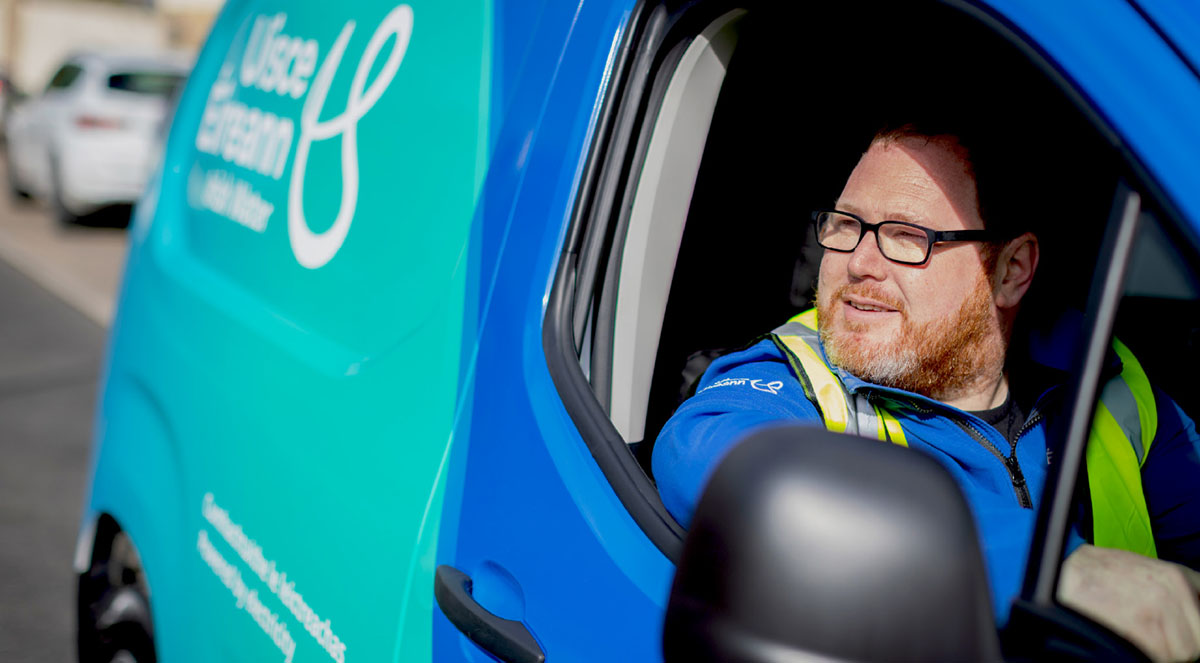 Tiománaí flít Uisce Éireann, ar a bhfuil meangadh gáire, suite i veain ar a bhfuil an branda