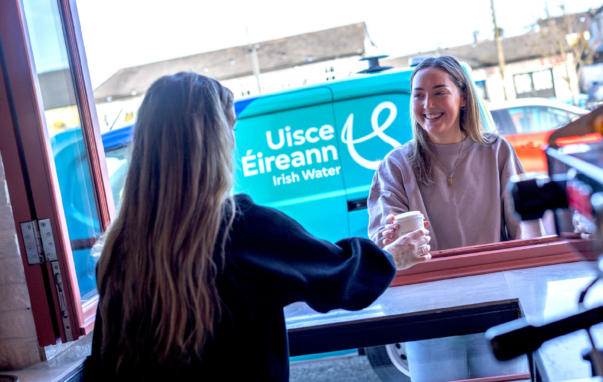 Barista serving a female customer through a hatch in the cafe with branded Uisce Eireann van in the background