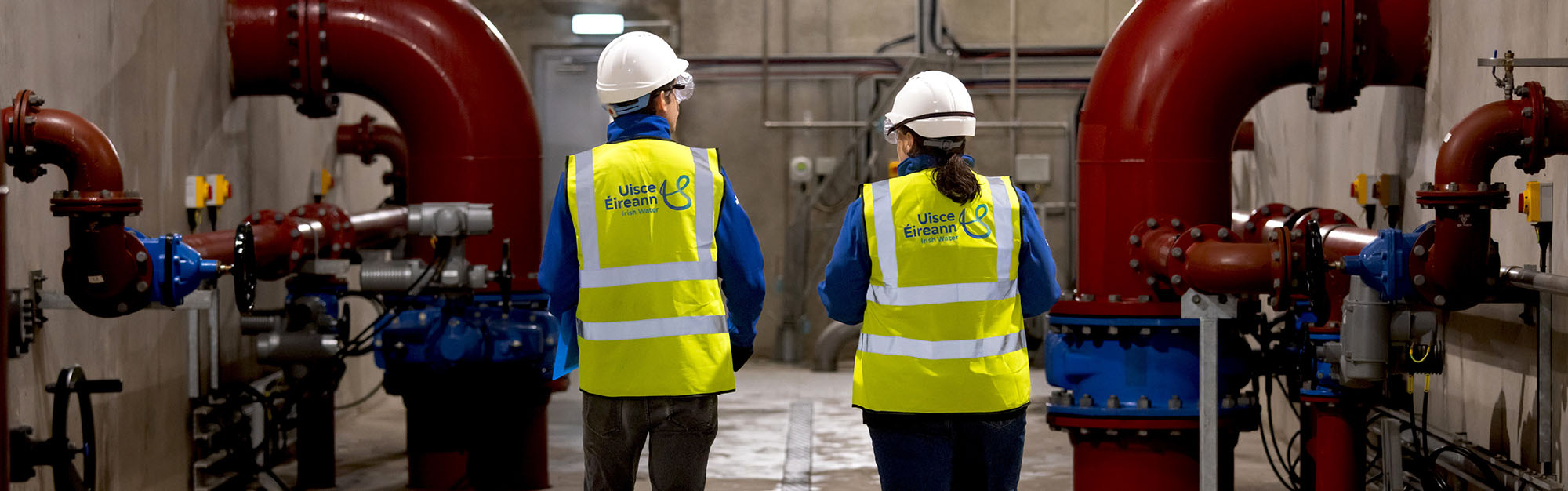 Male and female Uisce Eireann employees walking through Arklow Wastewater Treatment plant among large red pipes with valves and monitoring devices