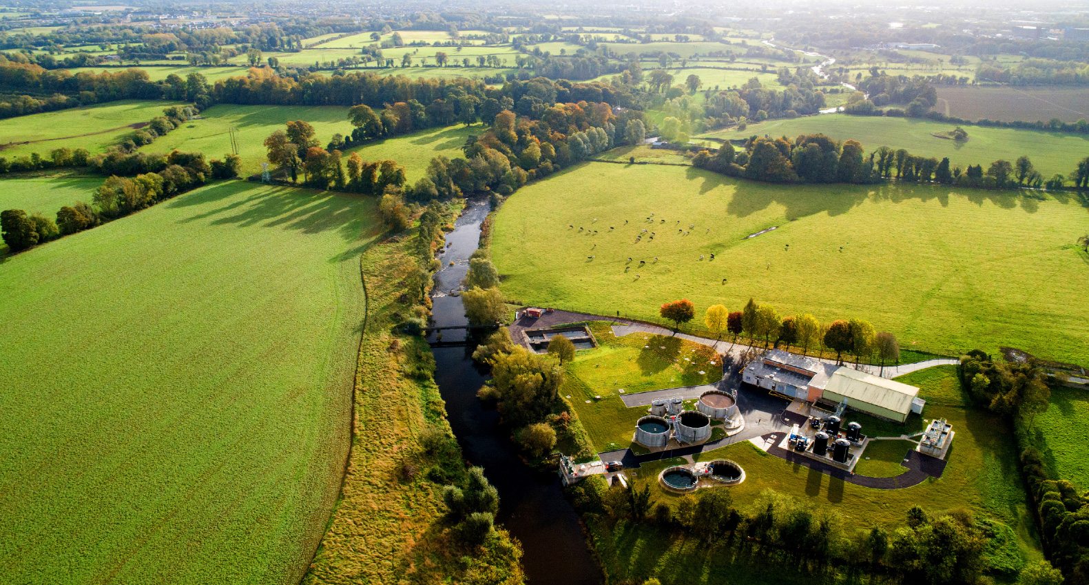Aerial view of a meandering river at the end of a water treamtment plant amongst a vast area of green fields in sunshine.