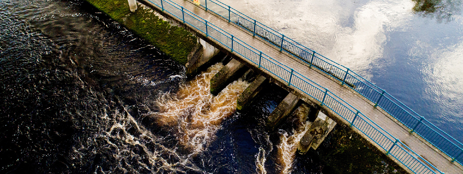 Aerial view of both sides of a dam as water gushed through. A walkway over the dam is evident.