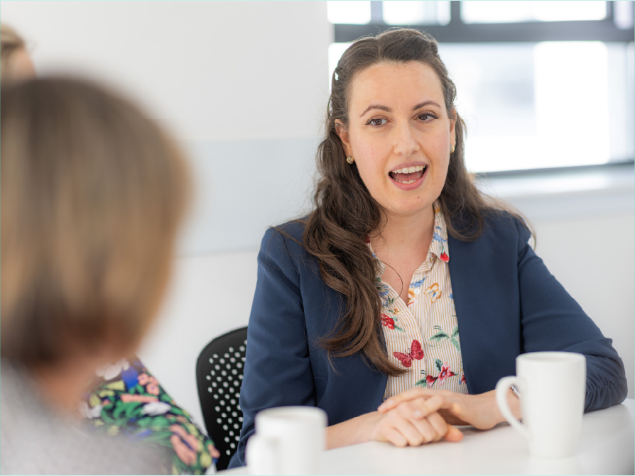 3 people in a meeting environment with mugs in front of them in a relaxed posture. Only one female is in focus.