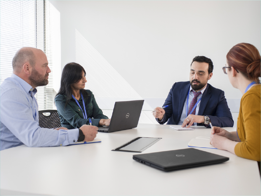 4 people of different gender with laptops in an office meeting setting taking notes as one person talks.