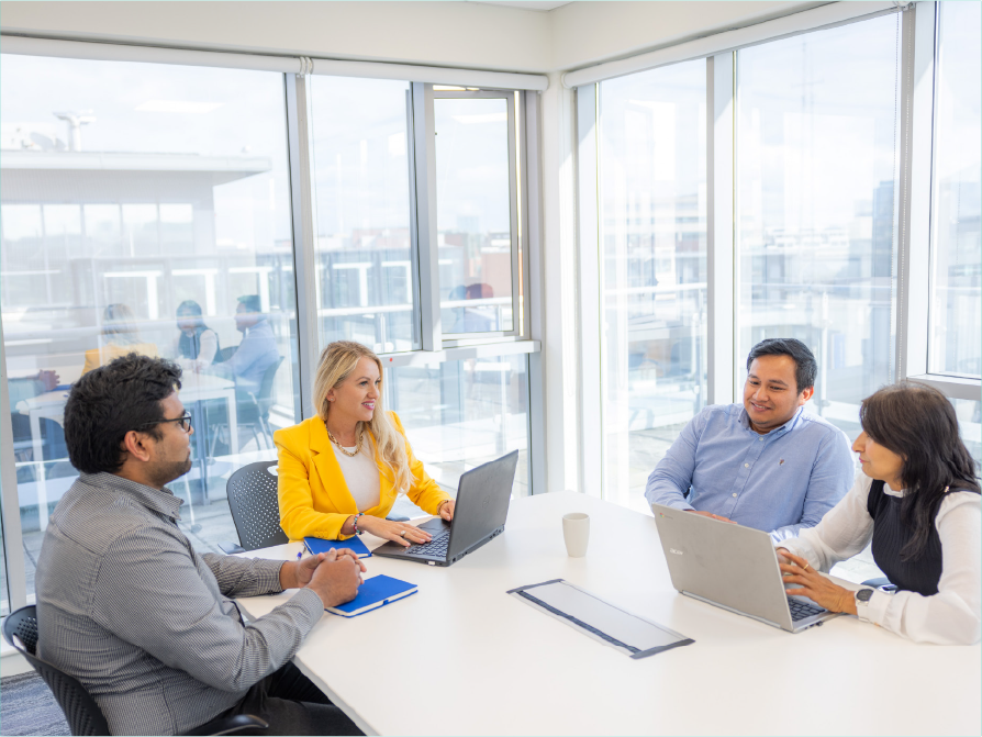 4 people in a bright office environment in a meeting setting with laptops and notepads.