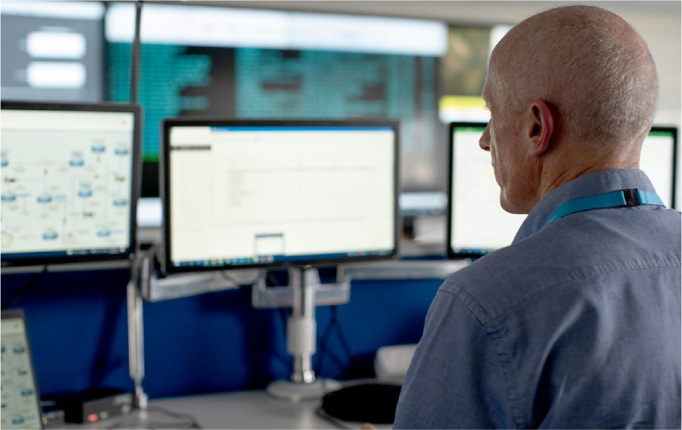 Technical operative in an office monitorig setting with numerous screens in his view and a large wall screen in the background.