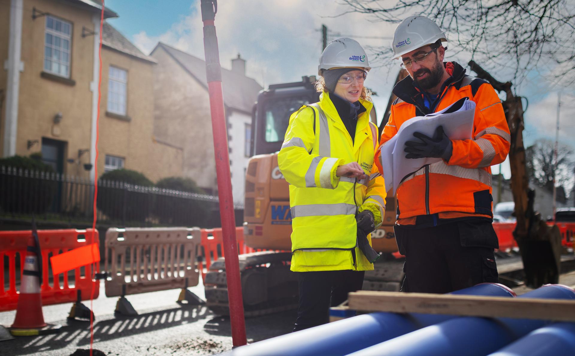 Two Uisce Éireann engineers doing road works with pipes looking at plans