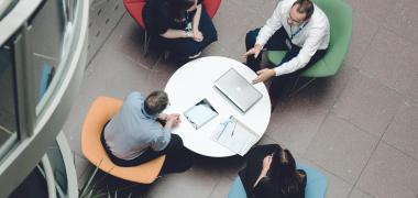 An aerial shot of four office workers sitting on chairs around a table in a meeting
