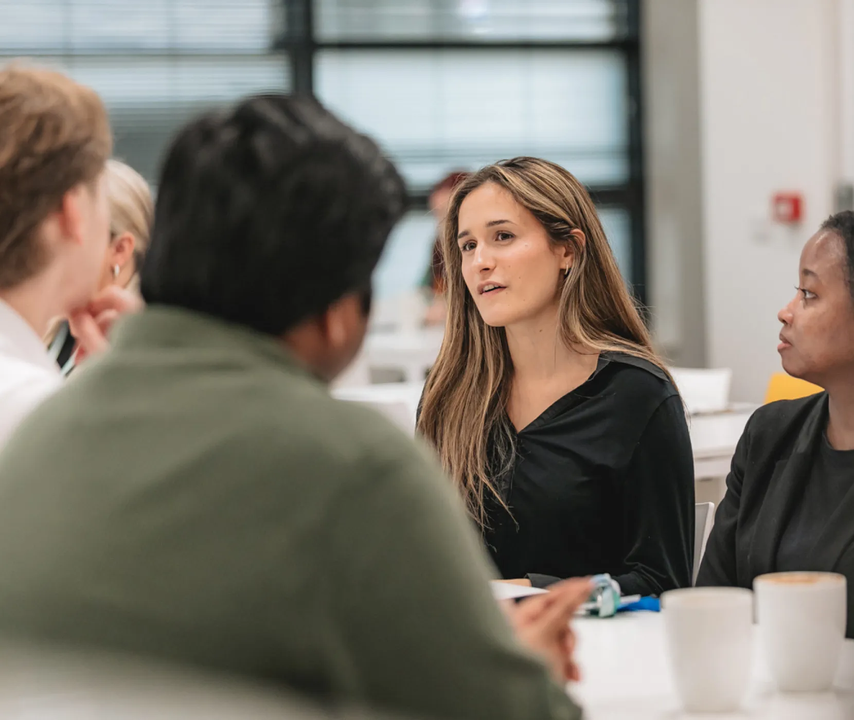 A lady speaking in front of two men on a table and another lady looking at her