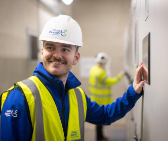 UÉ employee wearing a hard hat and smiling at the camera. His hand is holding a Ipad on the wall