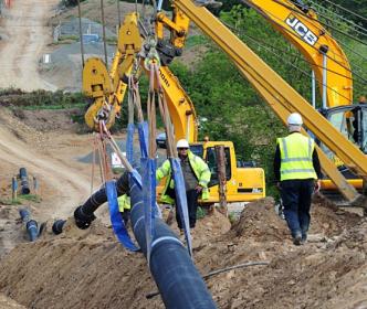 Two men walking through a construction site as a row of pipes are being lifted into place by cranes